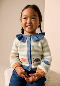 Gebreide cardigan met blauwe kraag en knopen, voorzien van pastelkleurige strepen en vissenpatronen in oranje en blauw, gecombineerd met blauwe spijkerbroek.