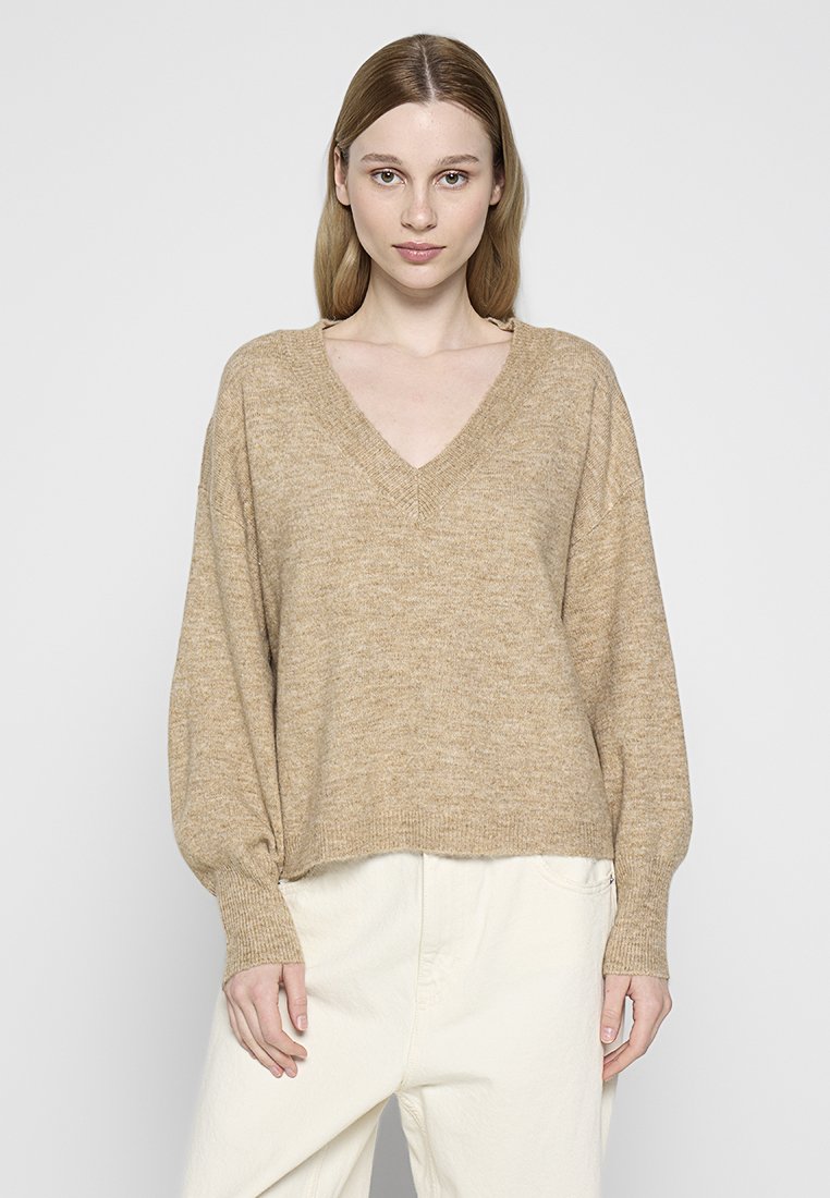 Suéter de punto beige con escote en V, mangas largas tipo globo y un corte holgado, combinado con pantalones de color claro. Textura suave y diseño casual.