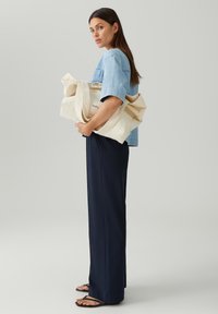 Canvas tote bag in crèmekleur met logo, gecombineerd met een lichtblauwe denim shirt en marineblauwe wijde broek. Model staat in profiel tegen een grijze achtergrond.