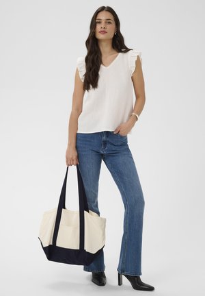 Frau in weißem ärmellosem Rüschen-Top und blauer Jeans, die eine große beige-schwarze Tragetasche hält und schwarze Stiefel trägt.