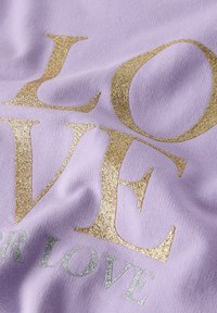 Tissu lilas avec de grandes lettres dorées scintillantes formant le mot "LOVE" et de plus petites lettres argentées pailletées en arrière-plan sur un textile doux.