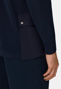 Prenda de punto color azul marino con un panel lateral que incluye un bolsillo con cierre de broche. Se pueden ver puños acanalados y un detalle de tela texturizada.