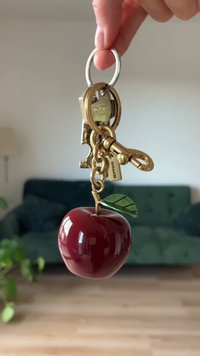Porte-clés en forme de pomme rouge avec une finition brillante, attaché à un porte-clés en métal doré et à une chaîne. Une touche de feuille verte est incluse.