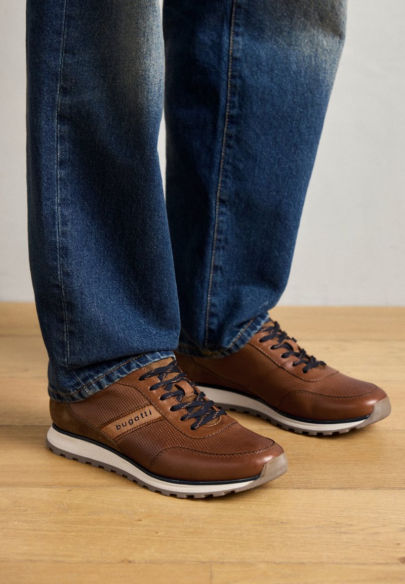 Baskets en cuir marron avec des panneaux latéraux texturés, semelle en caoutchouc blanche, lacets noirs et logo "bugatti" sur le côté, portées avec un jean bleu.