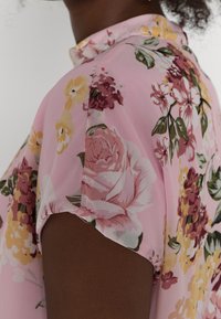 Blommig blus i rosa tyg med upprullade ärmar, med stora rosor och mindre blommor i gult och burgundy, samt gröna blad.