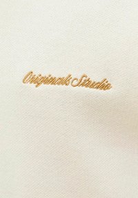 Krém színű anyag, amelyen egy arany színű, hímzett "Originals Studio" logó található, sima textúrával és finom varrási részletekkel.