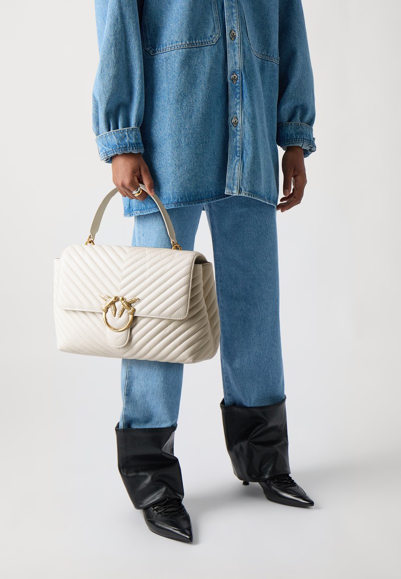 Kviltota krēma krāsas rokassoma ar zelta furnitūru, kurai ir apaļa sprādze. Valkāta ar džinsu krekla, gaiši zilu džinsu un melniem asiem zābakiem.