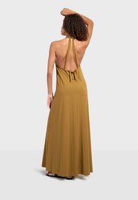 Femme aux cheveux bouclés portant une longue robe dos nu moutarde à col halter et des sandales noires, debout avec un bras levé derrière la tête.