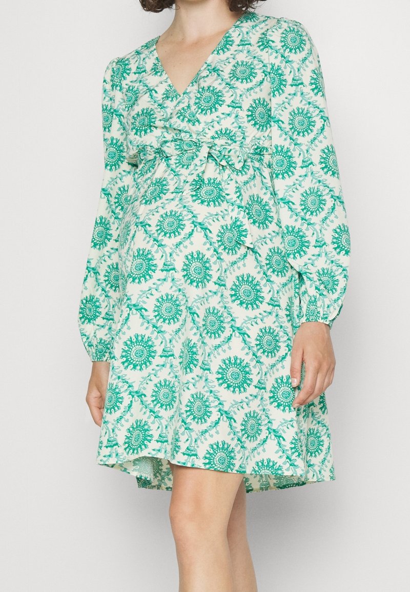 Femme portant une robe portefeuille mi-longue à manches longues, à motifs verts et blancs, avec un nœud ceinturant la taille.