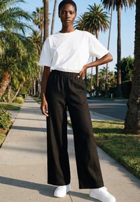 Giovane con capelli corti in piedi su un marciapiede fiancheggiato da palme, indossa una maglietta bianca semplice, pantaloni neri a gamba larga e sneakers bianche.