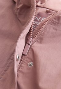 Gros plan d'une veste rose poudré partiellement dézippée avec les dents en plastique de la fermeture éclair visibles et un bouton-pression en métal.