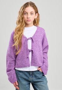 Cárdigan corto morado con cierre de lazo en el frente, mangas largas y tejido texturizado, usado sobre una camisa blanca y combinado con jeans de mezclilla.