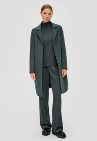 Groene wollen jas met een geknipt kraagje, gedragen over een donkere coltrui en bijpassende wijde broek. Dikke zolen op de schoenen.