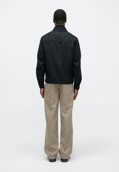 Vīrietis stāv ar muguru, ģērbies melnā jakā, bēšās platās biksēs un melnās kurpēs uz viendabīga baltā fona.