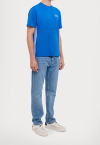 Blue crew neck t-shirt with white side stripes, paired with light blue jeans and light sneakers. Simple, casual design and fit.