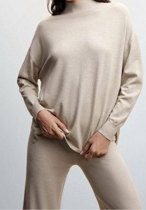 Femme portant un pull beige à col roulé à manches longues et un pantalon assorti, tenant l'ourlet inférieur du pull avec les deux mains.