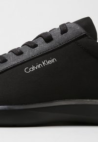 Schwarze Sneaker mit glattem Obermaterial aus Stoff und strukturierten schwarzen Schnürsenkeln. Verfügt über das weiße "Calvin Klein"-Logo an der Seite und eine elegante schwarze Sohle.