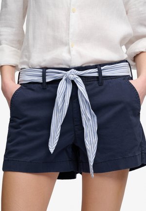 Persoon die marineblauwe shorts draagt met een blauw-wit gestreepte stoffen riem geknoopt in een strik en een wit overhemd met lange mouwen en knopen.
