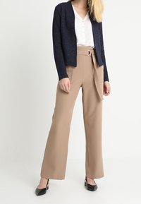 Námořnický texturovaný kardigan, bílá blůza s malými tečkami, béžové široké kalhoty s poutky na opasek a kovovou sponou, černé lodičky s podpatkem.