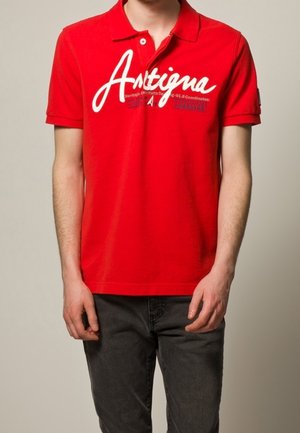 Punainen lyhythihainen poolopaita, jossa valkoinen "Antigua"-logo, kaulus ja nappilista, yhdistetty tummanharmaisiin farkkuihin.