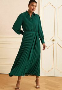 Abito midi plissettato verde con maniche lunghe, apertura a bottoni, collo a camicia e vita cintrata. Abbinato a tacchi dorati con cinturino.