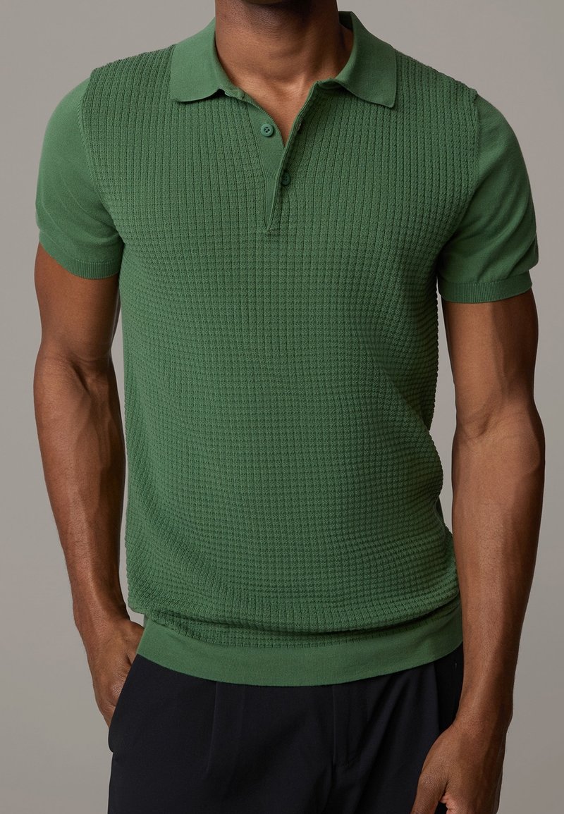 Homme portant un polo en maille vert près du corps à manches courtes avec un devant texturé et un pantalon noir, debout devant un fond uni.