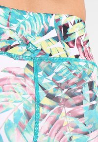 Zbliżenie kolorowego materiału w tropikalne liście z turkusowym przeszyciem i małą turkusową metką na pasku.