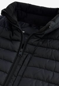 Chaqueta de pluma negra con cuello alto, cierre de cremallera y forro de forro polar en contraste. El material exterior está acolchado con una textura suave.