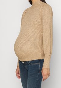 Beiger, gerippter Strickpullover mit hohem Kragen und gepolsterten Schultern, tailliert geschnitten, kombiniert mit dunkelblauen Jeans.