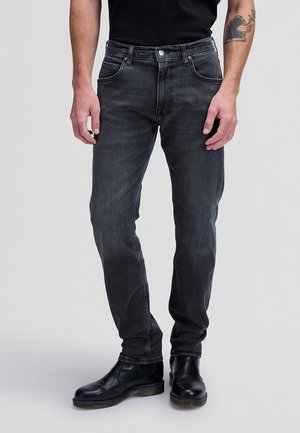Homme portant un jean noir slim délavé et des bottes en cuir noires, debout sur un fond clair.