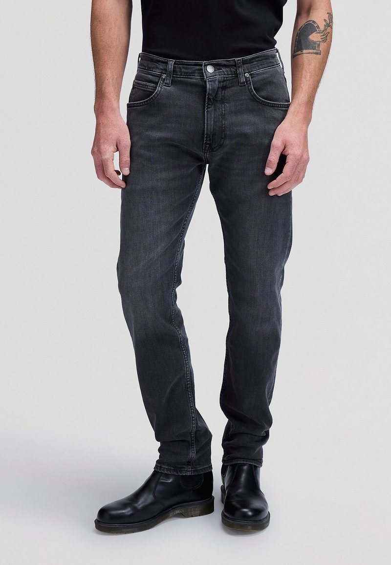 Homme portant un jean noir slim délavé et des bottes en cuir noires, debout sur un fond clair.