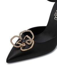 Scarpa nera in pelle con tacco alto e punta affusolata, con un dettagliato fiocco impreziosito da cristalli dorati sul davanti e una raffinata cinghia alla caviglia.