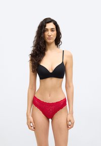 Γυναίκα με μακριά σγουρά σκούρα μαλλιά, ντυμένη με μαύρο σουτιέν και κόκκινα εσώρουχα από δαντέλα, στέκεται μπροστά σε απλό φωτεινό φόντο.
