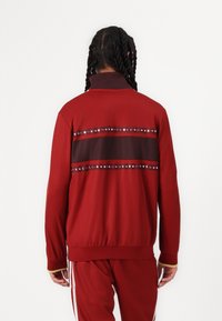 Röd träningsjacka med ett mörkt rand över ryggen, prydd med dekorativa nitar. Slät tyg, hög krage och ribbade muddarna.