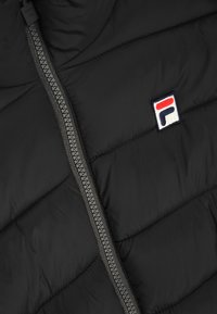 Giacca a vento nera con texture trapuntata, zip visibile grigia e un piccolo logo ricamato con colori rosso, bianco e blu.