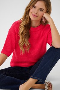 Heller rosa gestrickter Pullover mit rundem Hals und kurzen, umgeschlagenen Ärmeln. Getragen mit dunkelblauen Jeans. Die Textur wirkt leicht klobig.