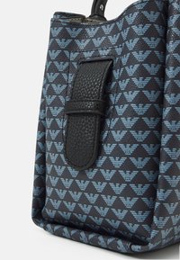 Ciemnoniebieska torba tote z wzorem, z powtarzającymi się jasnoniebieskimi logotypami orła oraz czarnym, teksturowanym skórzanym paskiem po boku.