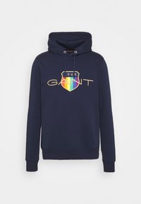 Sweat à capuche navy avec un logo frontal jaune et un blason en dégradé arc-en-ciel. Les cordons au cou et les poignets côtelés complètent le design.