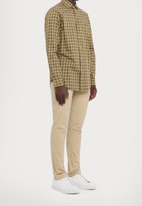 Man i en grön och beige rutig knäppskjorta, beigefärgade slim-fit byxor och vita sneakers, som står mot en enkel bakgrund.