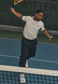 Tennise mängija valges lühikeste varrukatega särgis, millel on tekstureeritud muster, ja mustades spordipükstes, sooritab forehand lööki sinisel väljakul.
