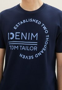 Námořnická modrá bavlněná tričko s kulatým grafickým motivem: "DENIM ZAŘÍZENÝ DVA TISÍCE SEVEN" ve světle modrém, tučném, bezpatkovém písmu.