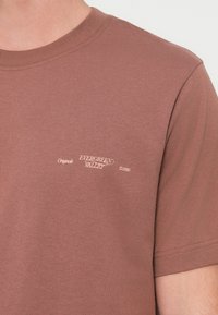 Καφέ βαμβακερό μπλουζάκι με στρογγυλή λαιμόκοψη, με λευκό σχέδιο που γράφει "Originals Evergreen Valley 1990" στην αριστερή πλευρά του στήθους.