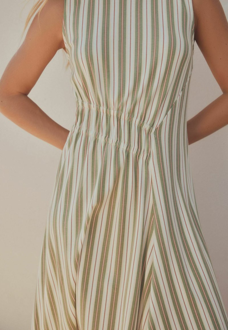 Robe sans manches à rayures verticales vertes, blanches et fines orange, taille élastiquée froncée, portée par une personne avec les bras derrière le dos.