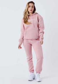 Sudadera rosa con capucha, bolsillo delantero y logotipo, combinada con pantalones jogger rosa a juego, ambos hechos de tela suave. Completado con zapatillas blancas para correr.