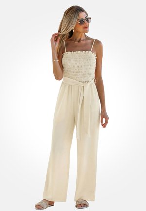 Kobieta w beżowym, bezrękawnym kombinezonie z marszczonym gorsetem, zawiązanym w pasie, w okularach przeciwsłonecznych i sandałach, dotykająca włosów i patrząca w prawo.