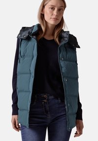 Teal pufferväst med dragkedja och tryckknappar, har huva och panelstickning. Buren över en mörkblå tröja och mörka jeans.