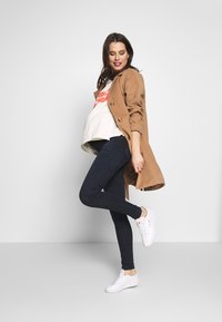 Casaco exterior bege com detalhes em botão, t-shirt gráfica branca, jeans skinny azul-escuro e ténis brancos. Em pose.