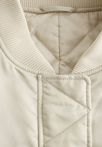 Beige quilted jacket with ribbed collar and reinforced stitching detail near the neckline and front closure.