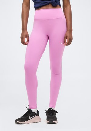 Kobieta nosząca różowe legginsy z wysokim stanem, czarno-różowe buty sportowe Nike oraz granatową, supełkowaną koszulkę na jednolitym tle.