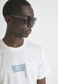 Witte t-shirt met een blauwe grafische print en tekst. De persoon draagt donkere sunglasses met een transparante grijze rand, die het ontwerp benadrukken.
